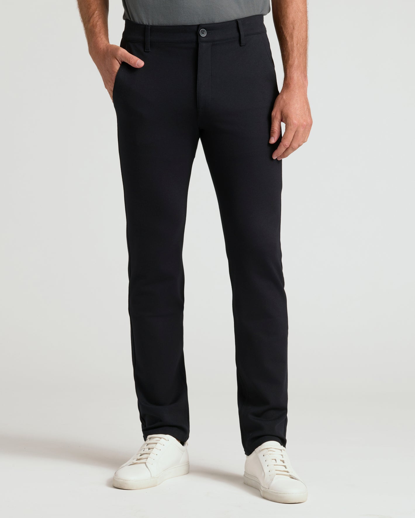 A person wearing slim-fit black trousers, paired with a gray top and white sneakers, standing against a light gray studio background.