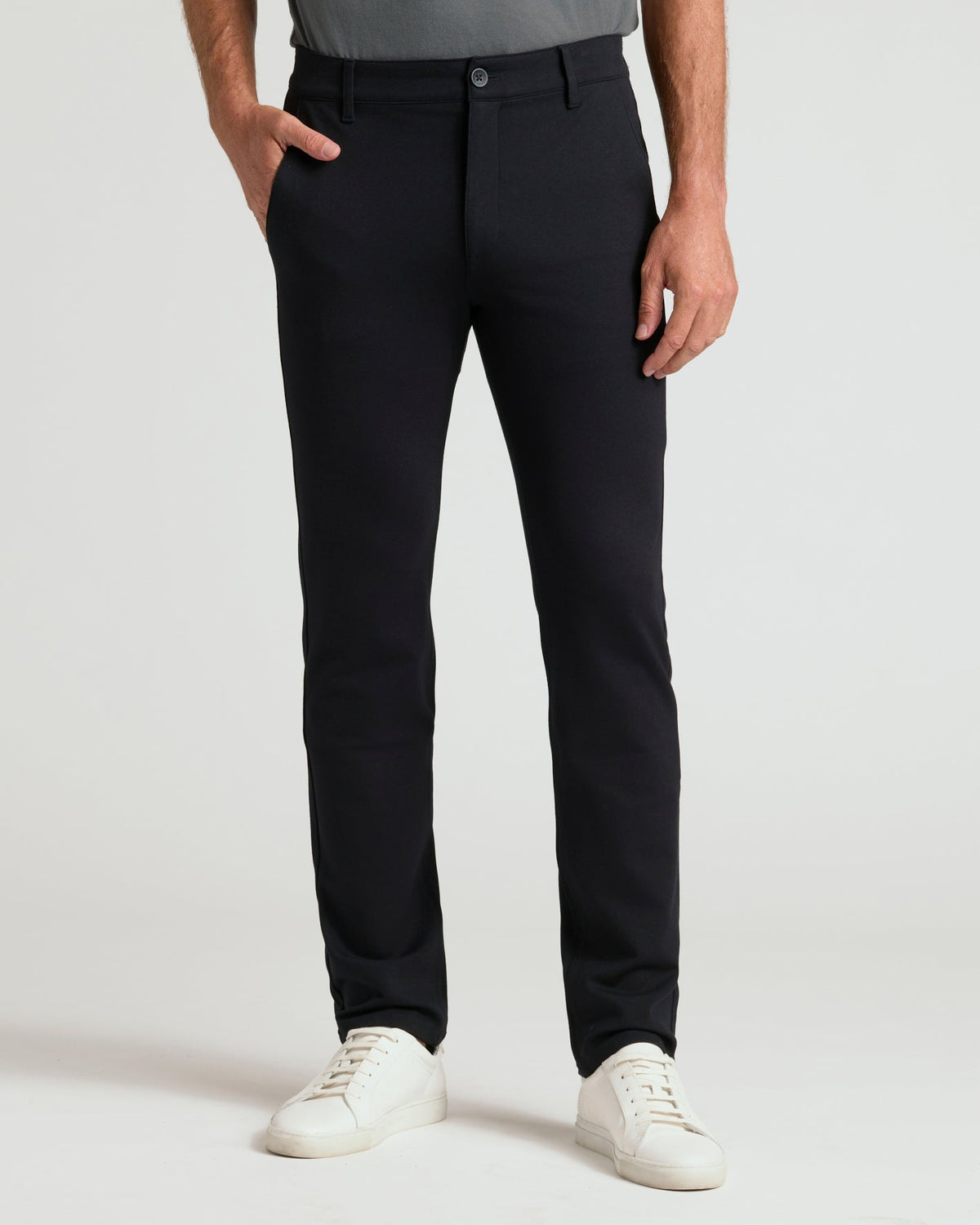 A person wearing slim-fit black trousers, paired with a gray top and white sneakers, standing against a light gray studio background.